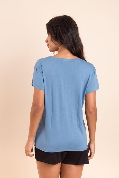 na foto, a modelo está de costas usando a t-shirt gola v na cor azul sereno, para evidenciar o comprimento da blusa